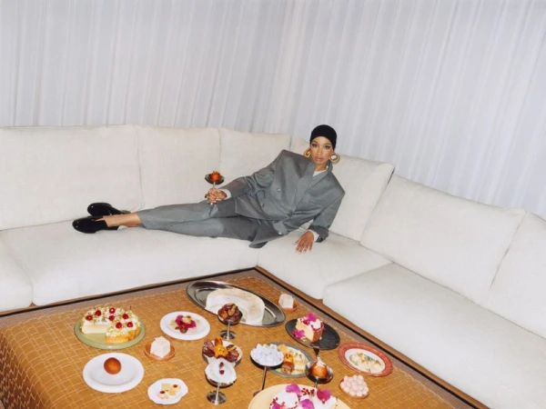 Model Ikram Abdi Omar reclining on a white sofa wearing a grey suit, with a spread of festive food on a table in front.