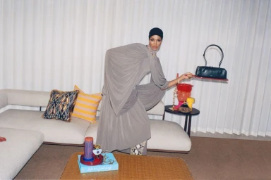 Model Ikram Abdi Omar wearing a grey draped dress holding a handbag in a living room for Selfridges