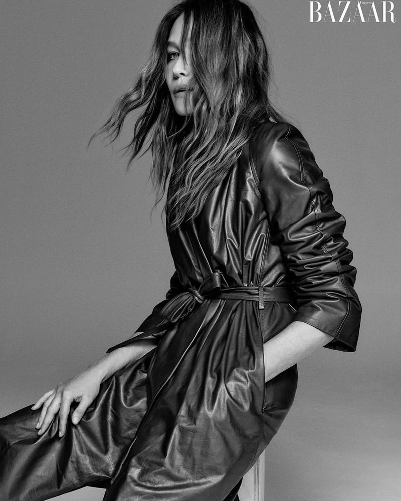 Monochrome portrait of a stylish adult woman in a black leather trench coat, sitting against a neutral backdrop with magazine logo.