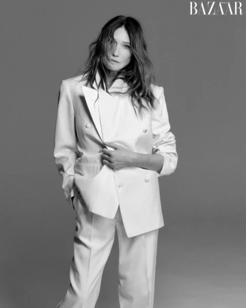 Carla Bruni wearing a white double-breasted suit holding her jacket lapel against a grey seamless background