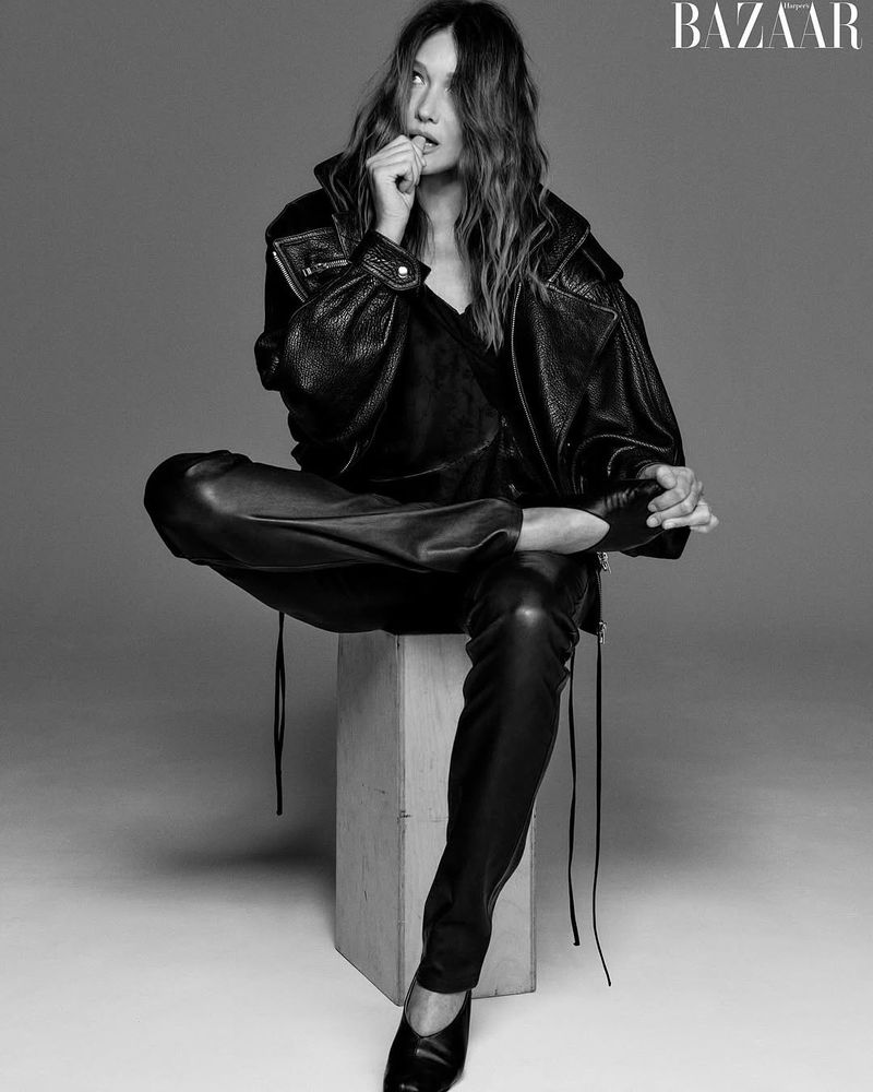 Black and white studio shot of an adult woman with long hair wearing a leather jacket and pants sitting on a box, fashion editorial.