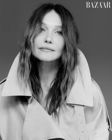 Monochrome portrait of Carla Bruni wearing a high-collared trench coat against a solid grey studio background.