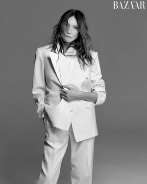 Carla Bruni wearing a white double-breasted suit holding her jacket lapel against a grey seamless background