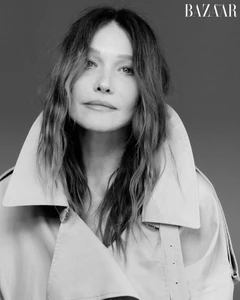 Monochrome portrait of Carla Bruni wearing a high-collared trench coat against a solid grey studio background.