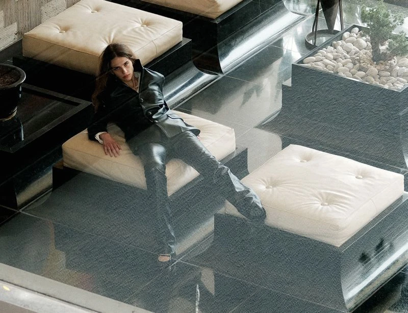 A stylish woman in a black leather jacket and pants lounges on a white cushion seat inside a modern indoor setting with black tiled floors.