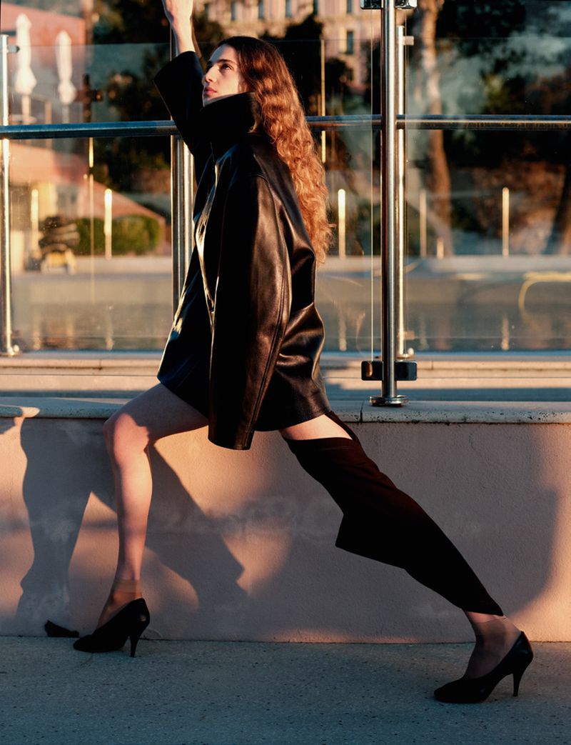 Model wearing a black jacket and heels posing outdoors by a glass railing during golden hour for Double Magazine editorial.