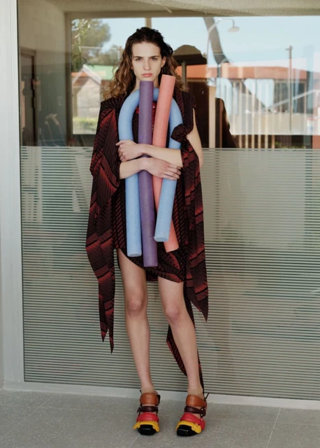 A person with long brown hair in a patterned brown outfit hugs three colorful pool noodles in front of a building with a modern design.