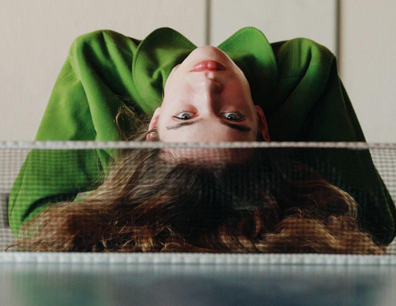 Portrait of a woman with long brown hair lying upside down in a green coat, reflected in a patterned glass table indoors.