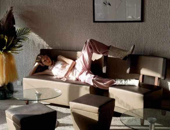 A woman reclines on a sofa indoors, dressed in pink, complemented by cream-colored boots, with a glass table and plant nearby.