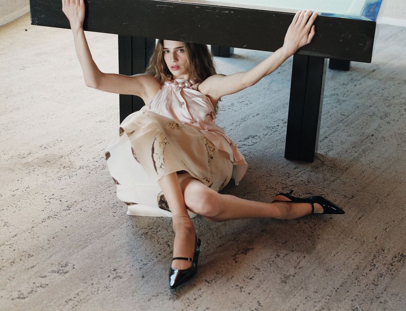 Young woman in a light pink dress sits beneath a black table, gazing at the camera with arms extended upwards, wearing black high heels.