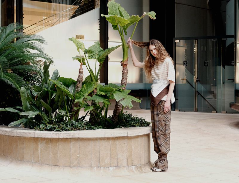 A woman in patterned pants stands near lush potted plants in a bright, modern indoor space with reflective glass and tile flooring.