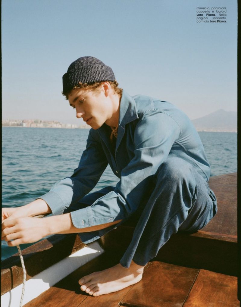 A fair-skinned male model in a blue denim outfit and knitted hat sits on a wooden boat against a sea backdrop with a distant cityscape.
