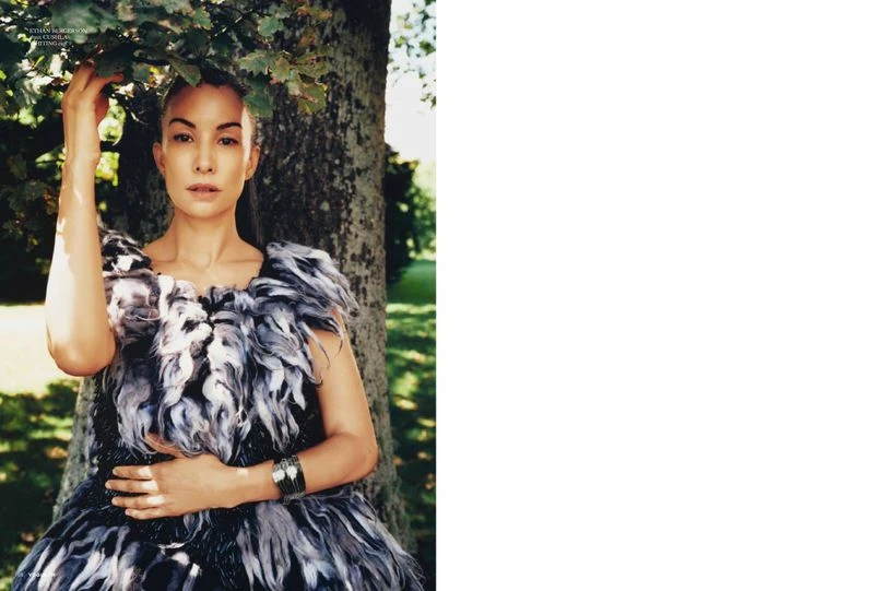 A woman in an elaborate, feathered dress stands beside a tree in a sunlit park setting, creating a natural and fashionable outdoor portrait.