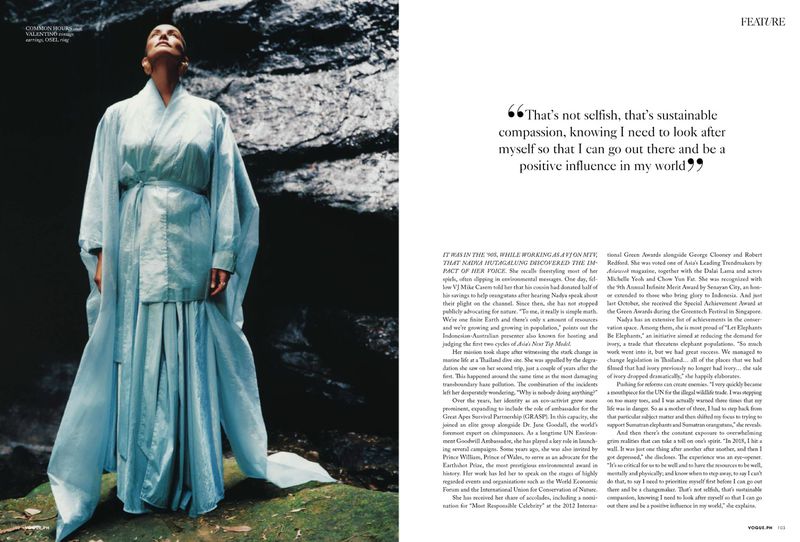 Fashion editorial shot of an adult woman in flowing light-blue dress standing outdoors against a dark rocky backdrop. Looking up.