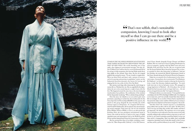 Fashion editorial shot of an adult woman in flowing light-blue dress standing outdoors against a dark rocky backdrop. Looking up.