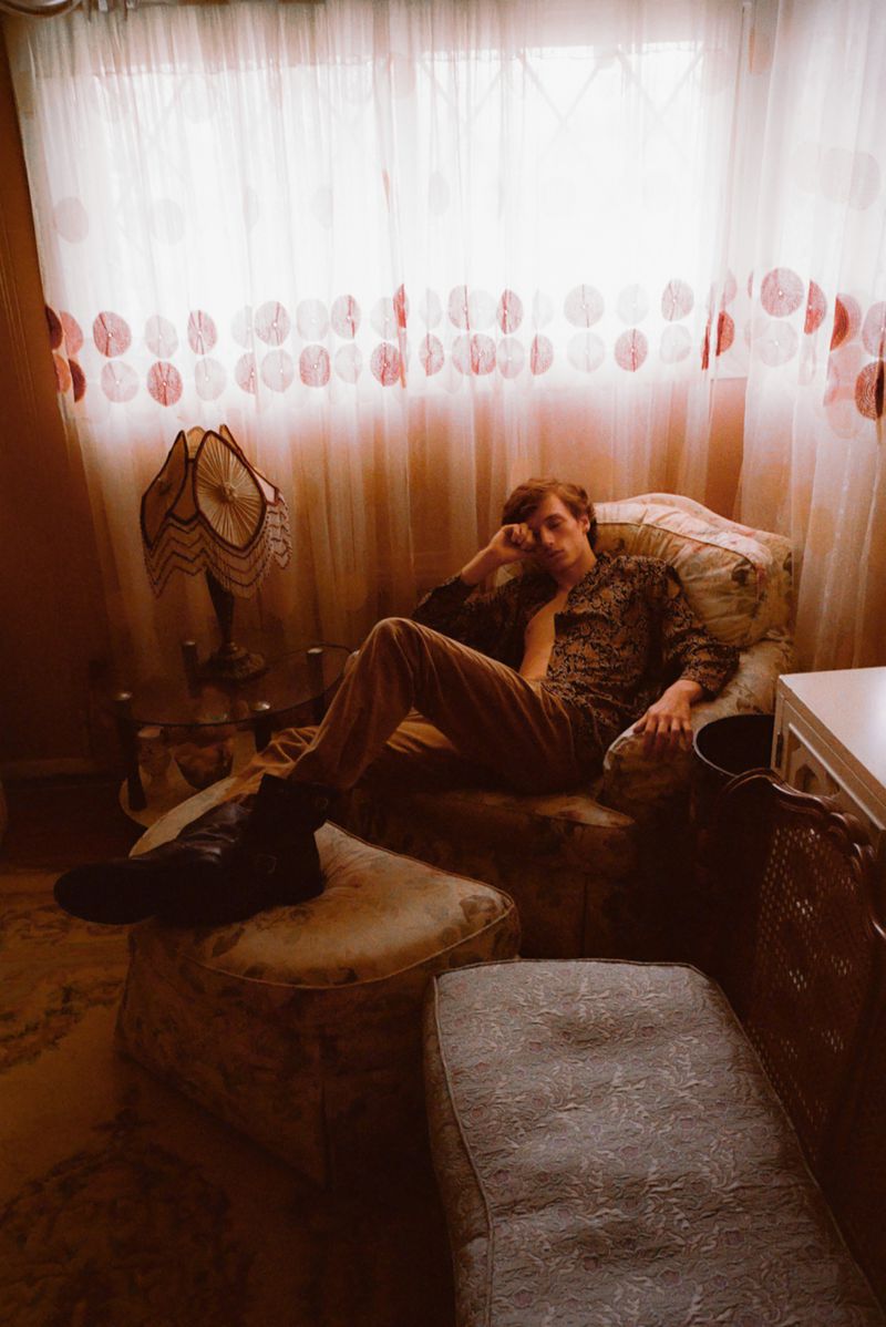 Young man relaxing in an armchair next to an ornate lamp and white patterned curtains, wearing a patterned jacket and trousers.