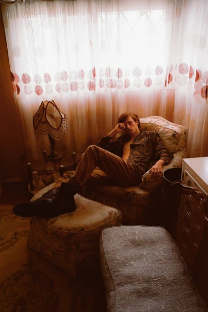 Young man relaxing in an armchair next to an ornate lamp and white patterned curtains, wearing a patterned jacket and trousers.