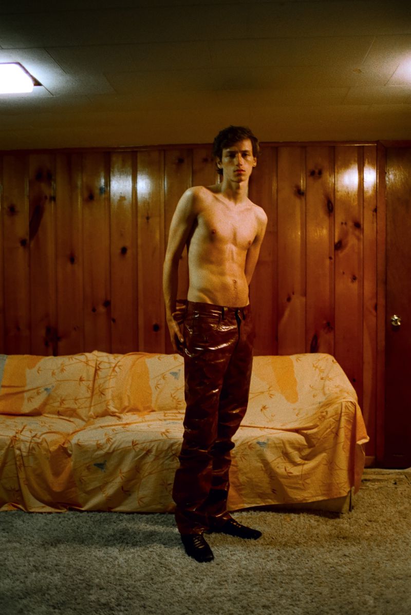 A slender, shirtless adult male poses in shiny maroon pants in an indoor setting with wood paneling and a beige couch; low-key lighting.