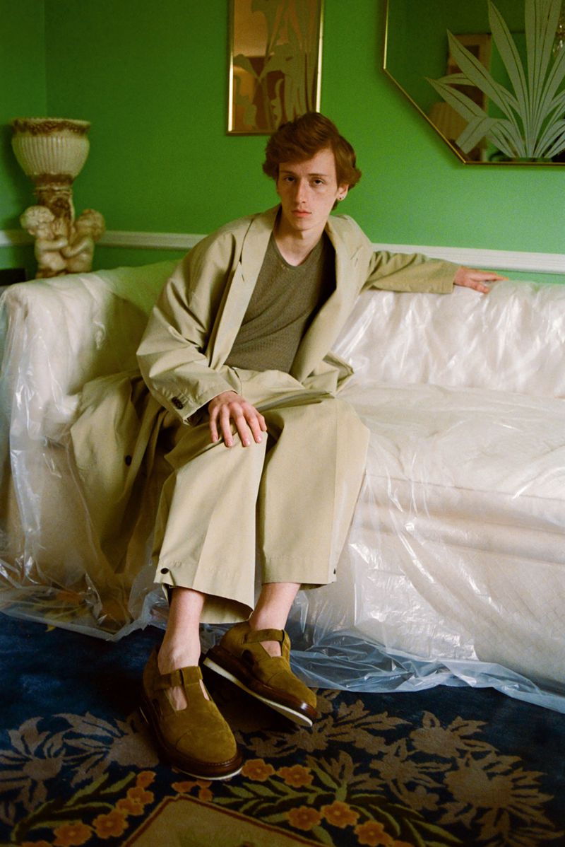 A red-haired male model sits on a plastic-covered couch in a green room, wearing a tan trench coat and brown shoes with a floral rug.