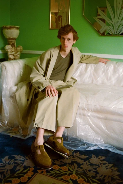 A red-haired male model sits on a plastic-covered couch in a green room, wearing a tan trench coat and brown shoes with a floral rug.