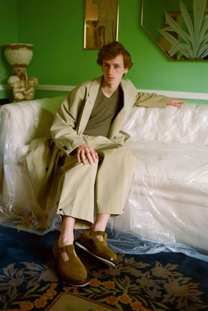 A red-haired male model sits on a plastic-covered couch in a green room, wearing a tan trench coat and brown shoes with a floral rug.