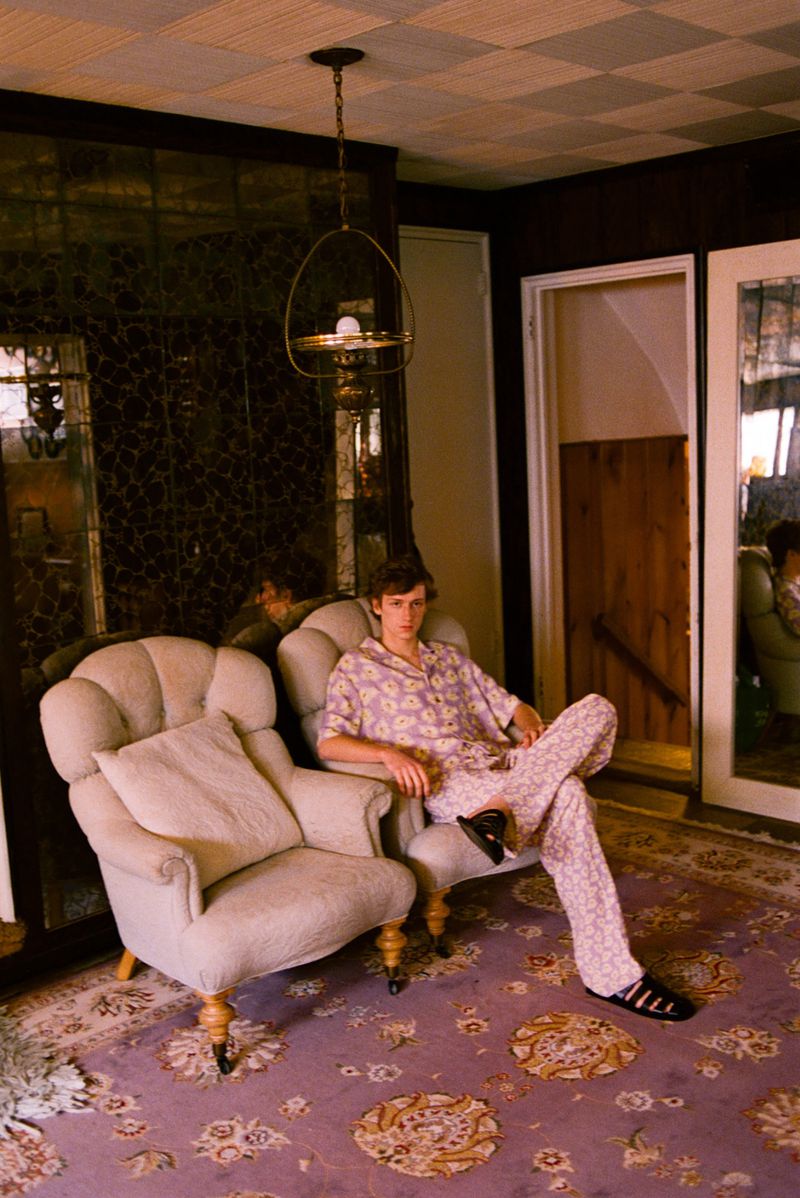 Man in patterned shirt and pants seated between two upholstered chairs on a patterned rug, beneath a hanging light fixture.