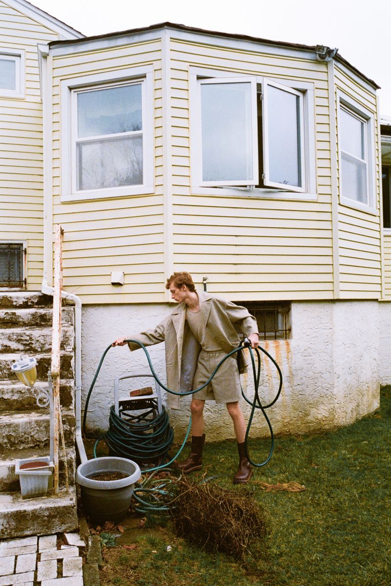 Young adult watering a backyard garden with a hose in a casual, neutral-toned outfit next to a yellow house with white-framed windows.