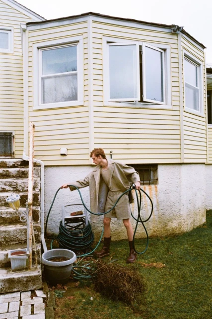Young adult watering a backyard garden with a hose in a casual, neutral-toned outfit next to a yellow house with white-framed windows.