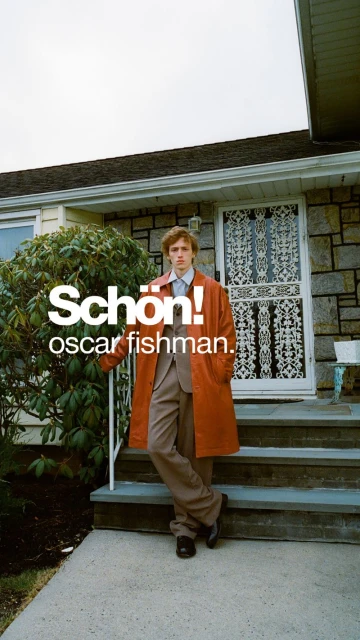 An adult male fashion model poses in front of a house, wearing a suit and orange trench coat, for an outdoor editorial fashion shot.
