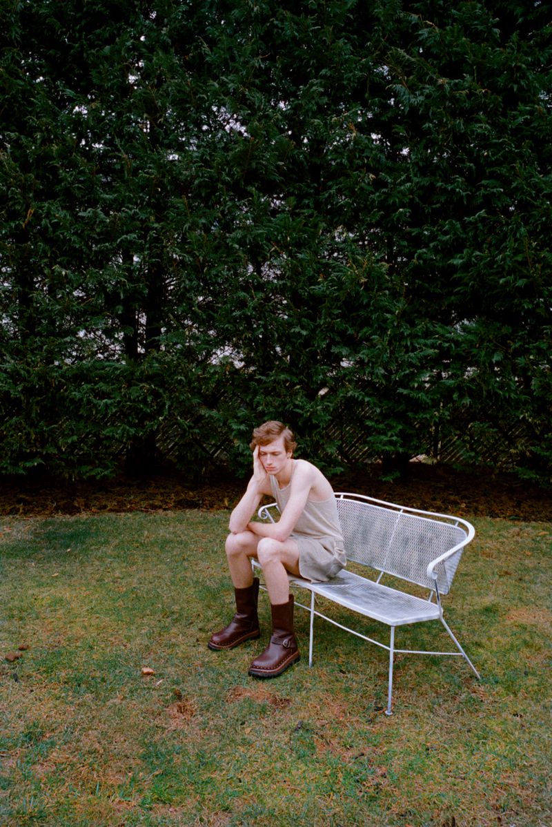 Young man sitting on a park bench in a yard with a large evergreen tree in the background wearing boots, shorts, and tank top.