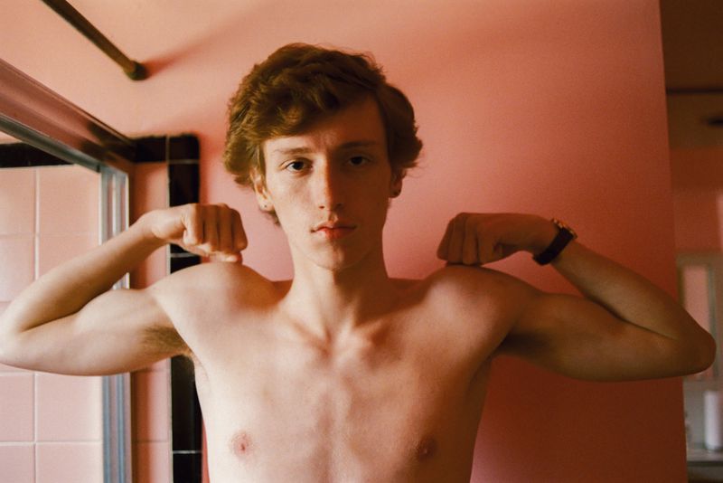 Young adult flexing muscles in an indoor setting, showcasing strength and fitness, with a pink tiled bathroom visible in the background.