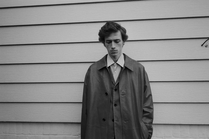 Black and white portrait of an adult male wearing a vest and overcoat, standing in front of a wall with horizontal siding.
