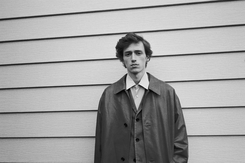 Monochrome portrait of a young man in a suit and overcoat, standing against a horizontal-panel wall in an outdoor setting with natural light.