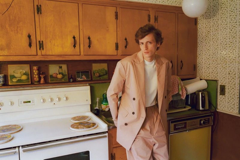 Young adult male in a pink suit stands in a retro kitchen with wood cabinets, vintage wallpaper, and an oven with a light green door.