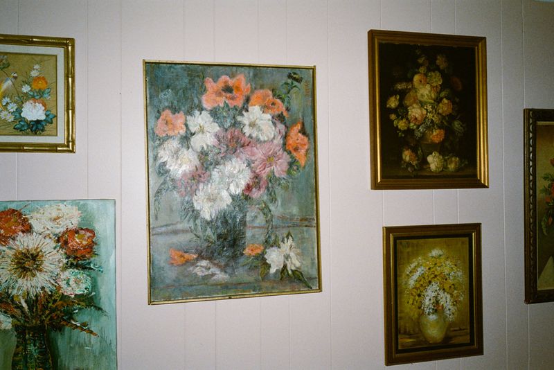 Collection of floral paintings in gold frames on a pastel pink wall, featuring arrangements of roses, daisies, and mixed blooms.