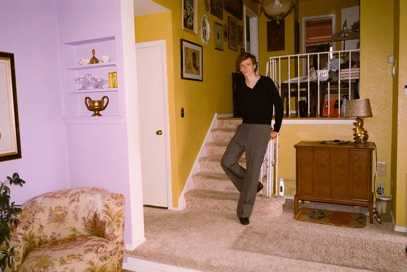 Indoor portrait of a slim adult man in a black shirt and grey trousers standing on carpeted stairs with patterned wallpaper in a home interior.