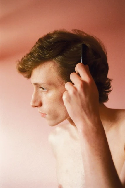 A young adult male is seen in a medium studio shot, combing his reddish-brown hair against a peach-colored backdrop.