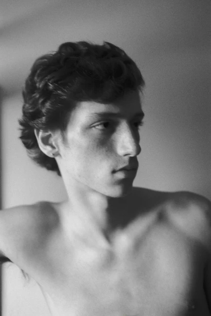 Monochrome portrait of a young adult male with dark curly hair, glancing to the side in a close-up shot against a neutral background.