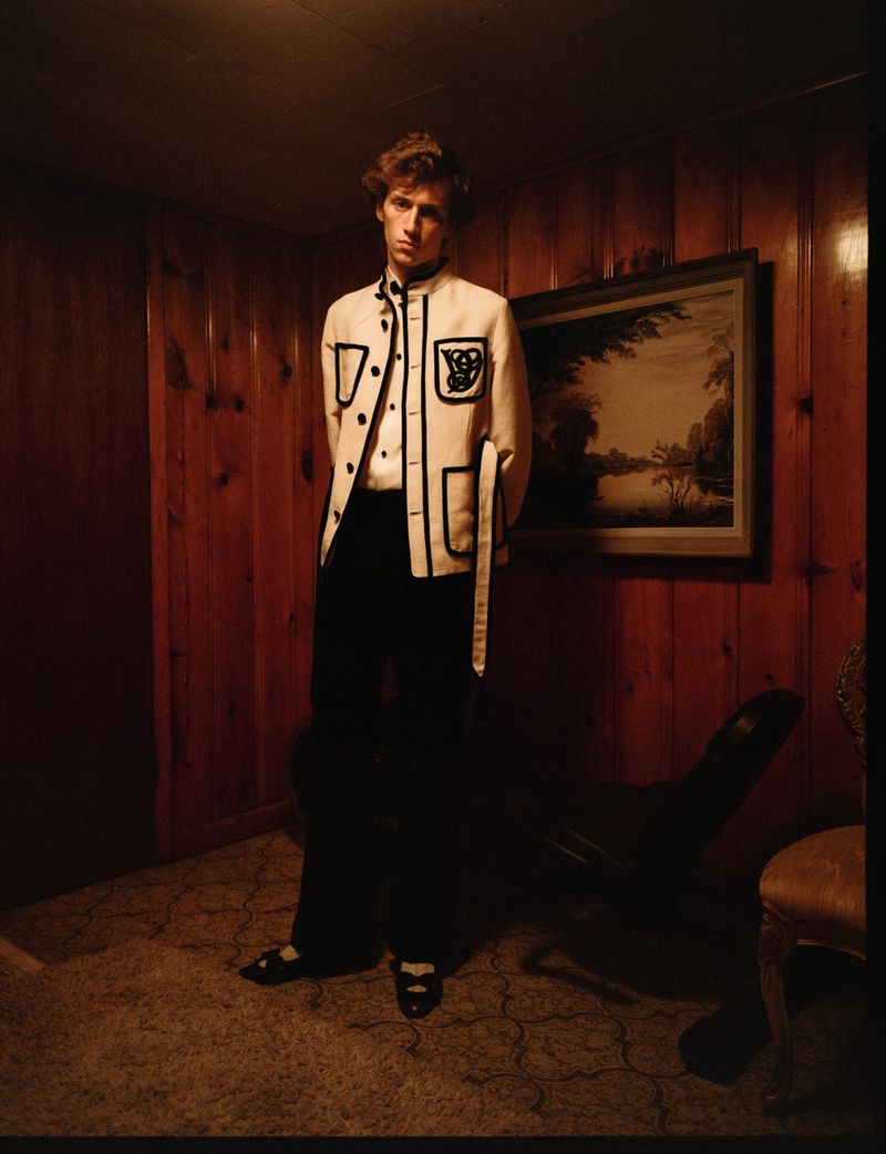 Male fashion model poses in a stylish cream-colored jacket with black accents in an indoor photo studio setting.