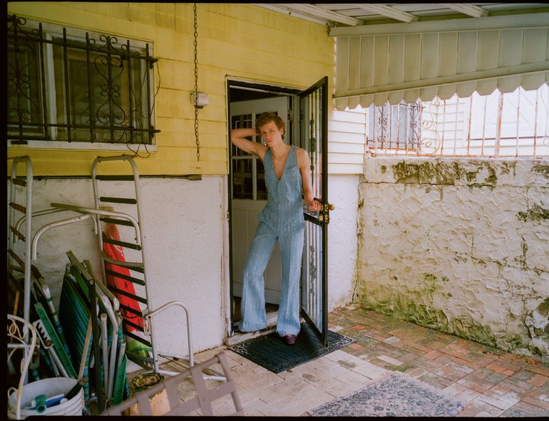 Woman in a light blue jumpsuit stands in a doorway leading to a yellow house with an outdoor patio area with a brick floor.