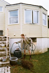 A young man in a beige coat and shorts holding a green garden hose in front of a yellow house for Schon Magazine