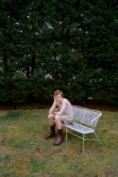 Young man sitting on a metal bench wearing a tank top, shorts, and dark boots in front of tall dark green trees.