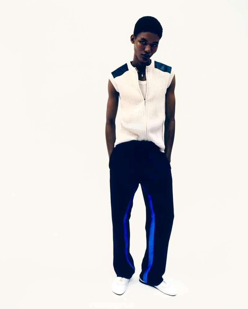 Fashion portrait of a black male model in a sleeveless white ribbed top and black pants with a blue stripe, set against a white background.