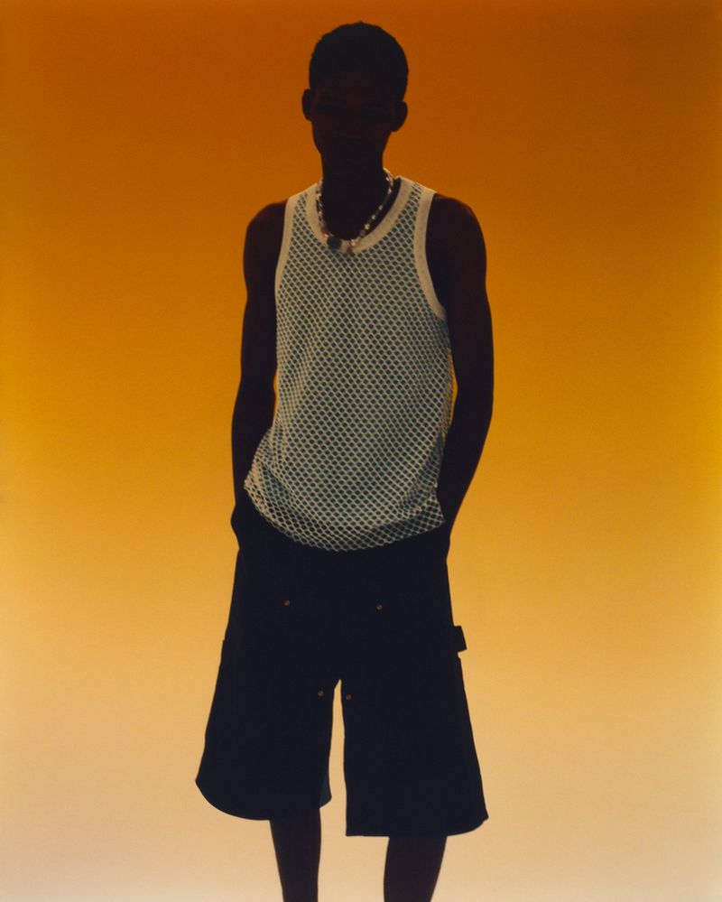 Dark silhouette of a man in a net tank top and baggy shorts against a gradient orange background, studio lighting. Fashion pose.