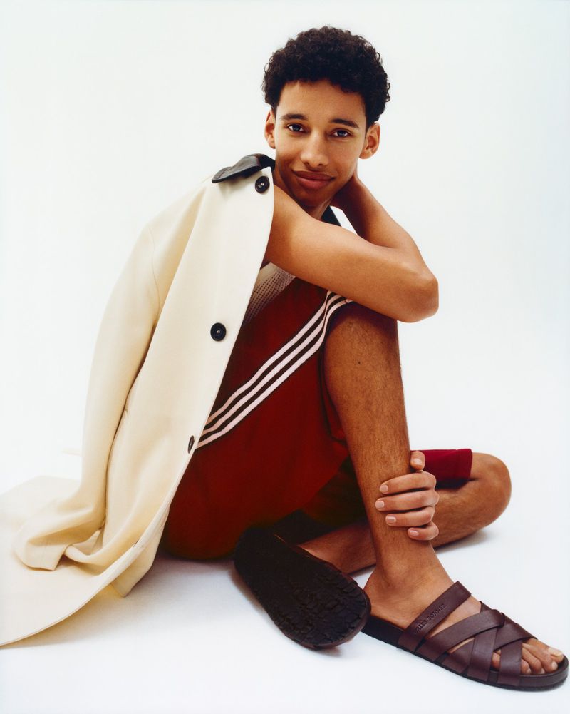 Young man with curly hair poses seated indoors wearing casual red shorts, brown sandals, and a beige jacket on his shoulder.