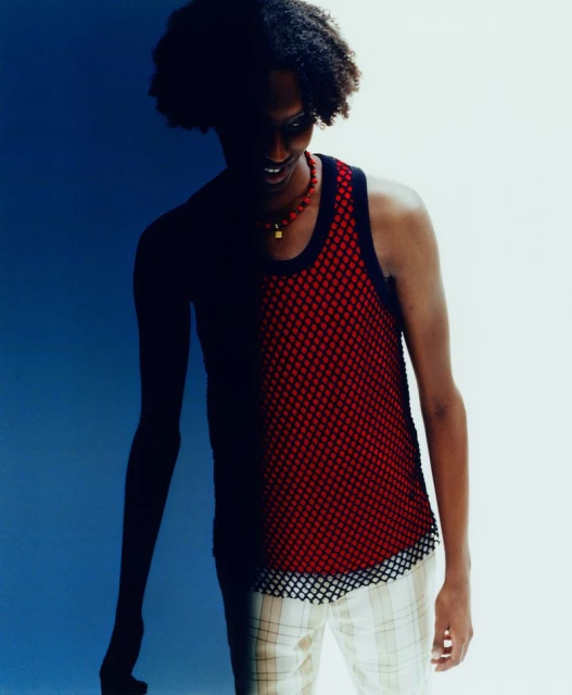 An adult stands indoors wearing a red mesh tank top and patterned trousers, in a medium shot with mixed lighting, against a blue and white background.