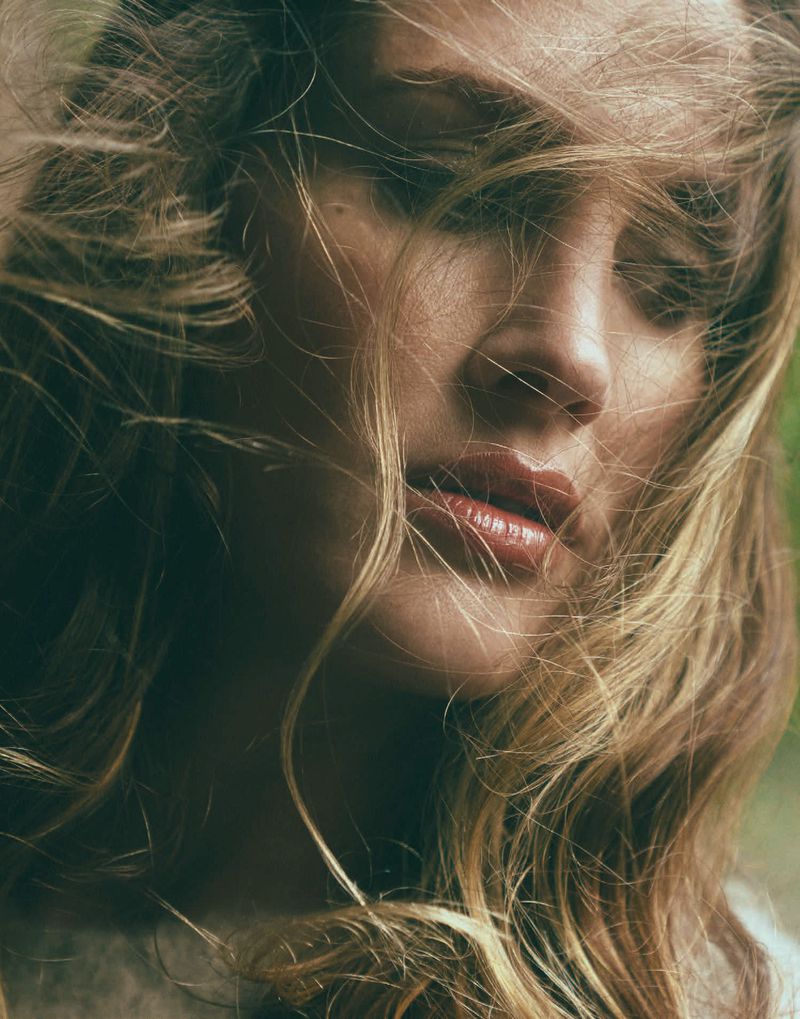 Close-up portrait of a woman with long blonde hair blowing across her face against a soft green background.