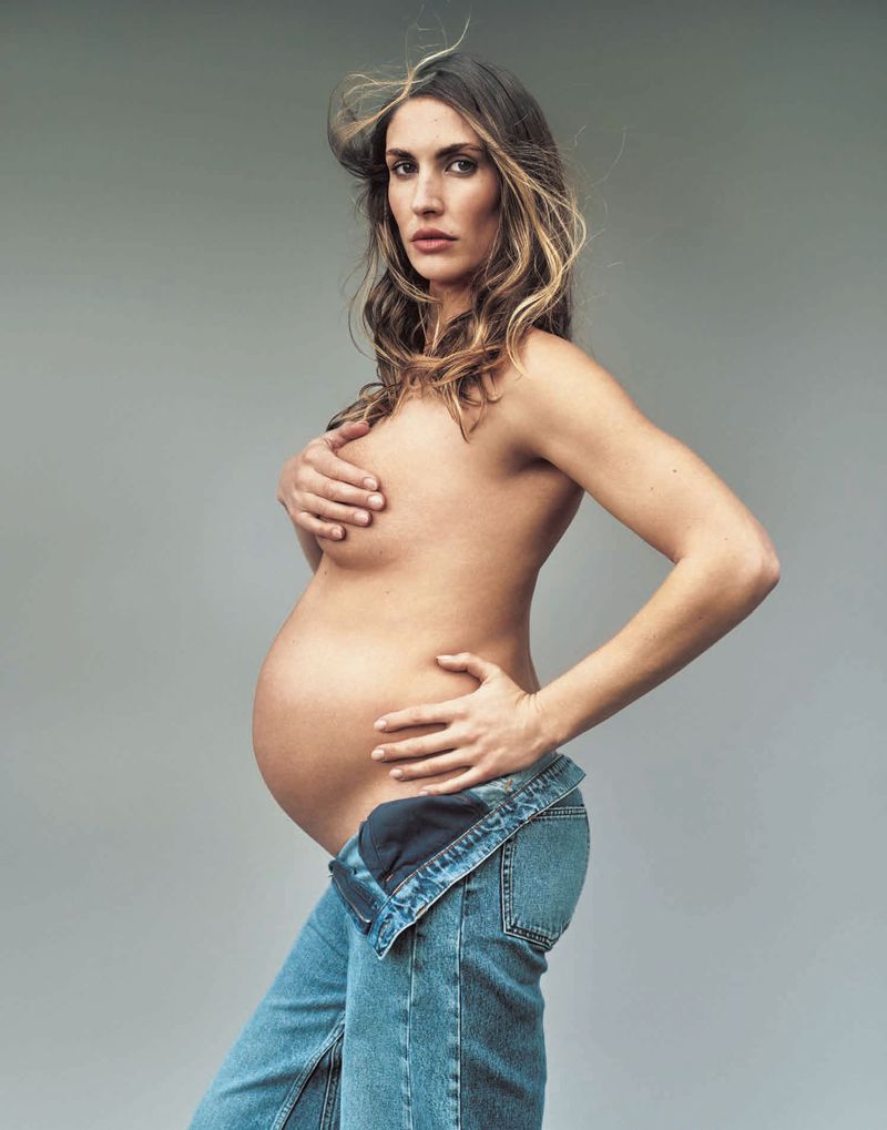 Pregnant woman in denim overalls poses in a studio environment with soft lighting, showcasing her bare torso and baby bump.
