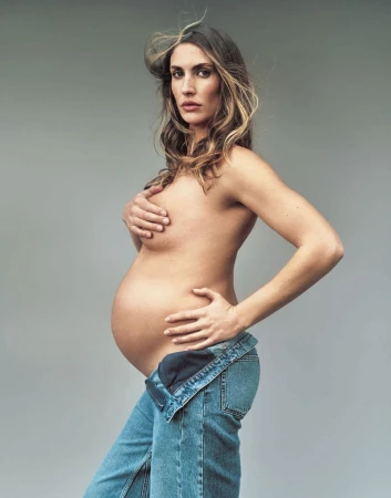 Pregnant woman in denim overalls poses in a studio environment with soft lighting, showcasing her bare torso and baby bump.
