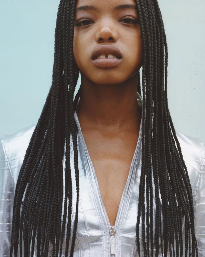 Portrait of a young black woman with long braids and a silver jacket against a light blue background in a studio setting.